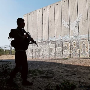 Hebräische Schrift auf einer Mauer, davor steht ein bewaffneter Mensch