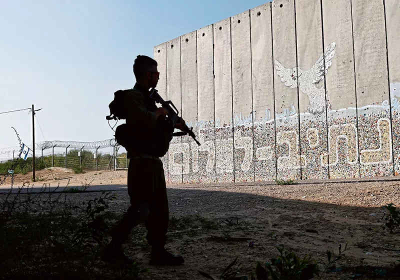 Hebräische Schrift auf einer Mauer, davor steht ein bewaffneter Mensch