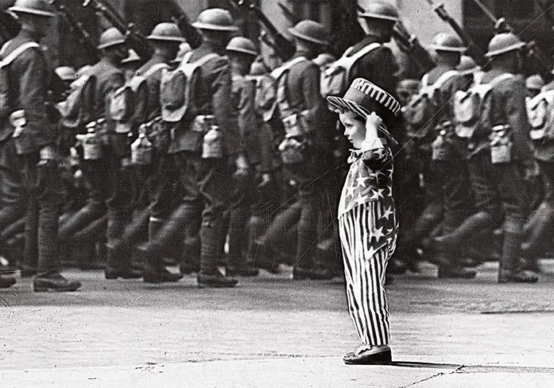 Schwarz-weiß Bild einer Militärparade mit einem salutierenden Kind im Vordergrund, das in eine US-Flagge gekleidet ist